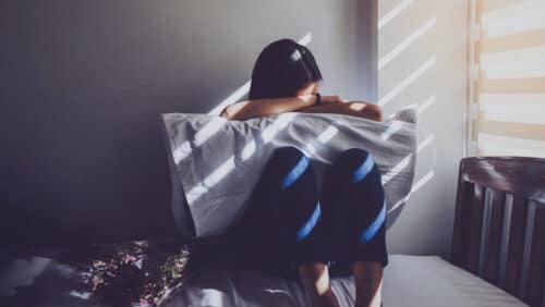 Asian women are sitting hugging their knees in bed. Feeling sad, disappointed in love in the dark bedroom and sunlight from the window through the blinds. Vintage tone.