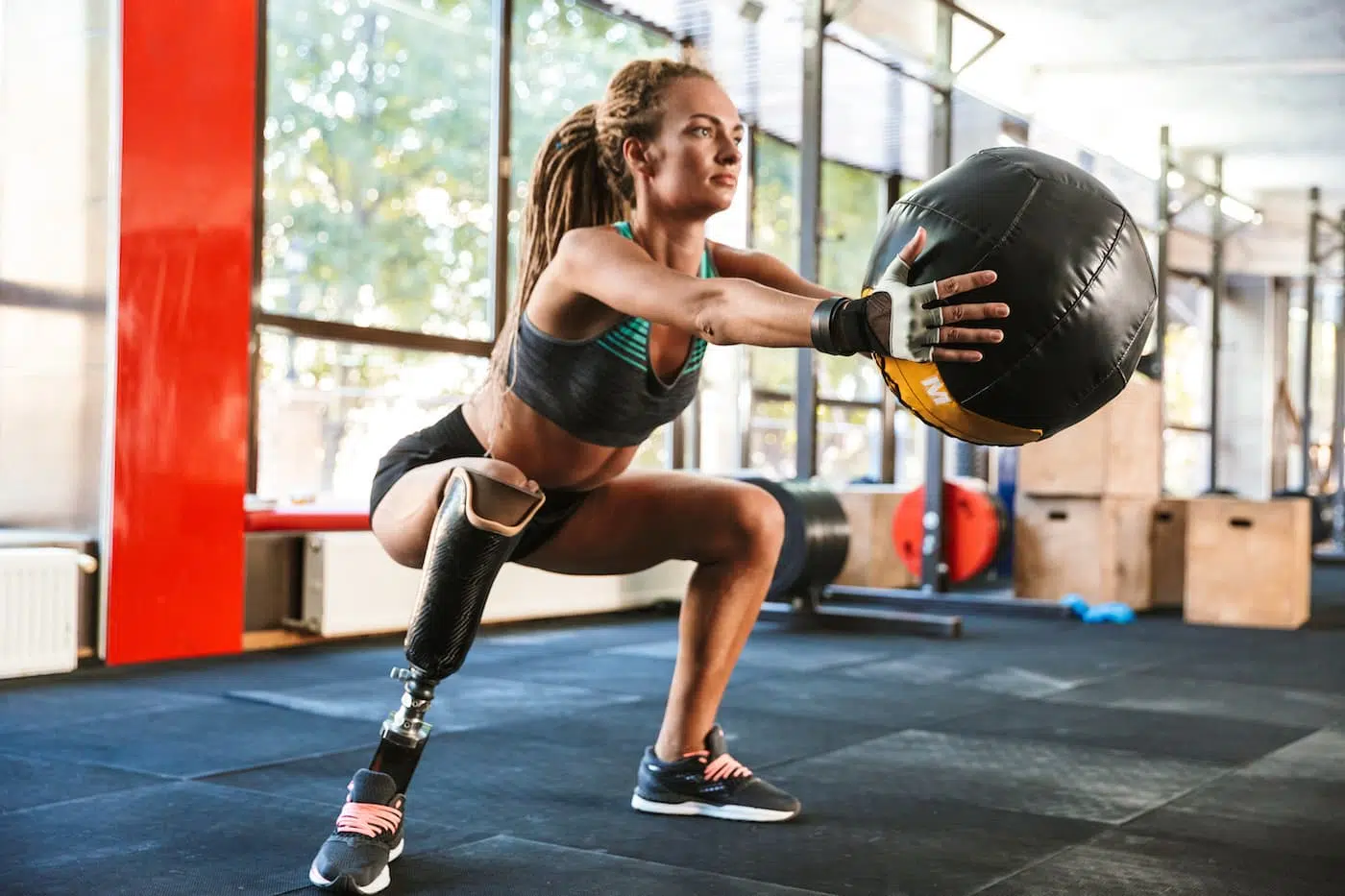 Woman with prosthesis in tracksuit doing squats with fitness ball in gym