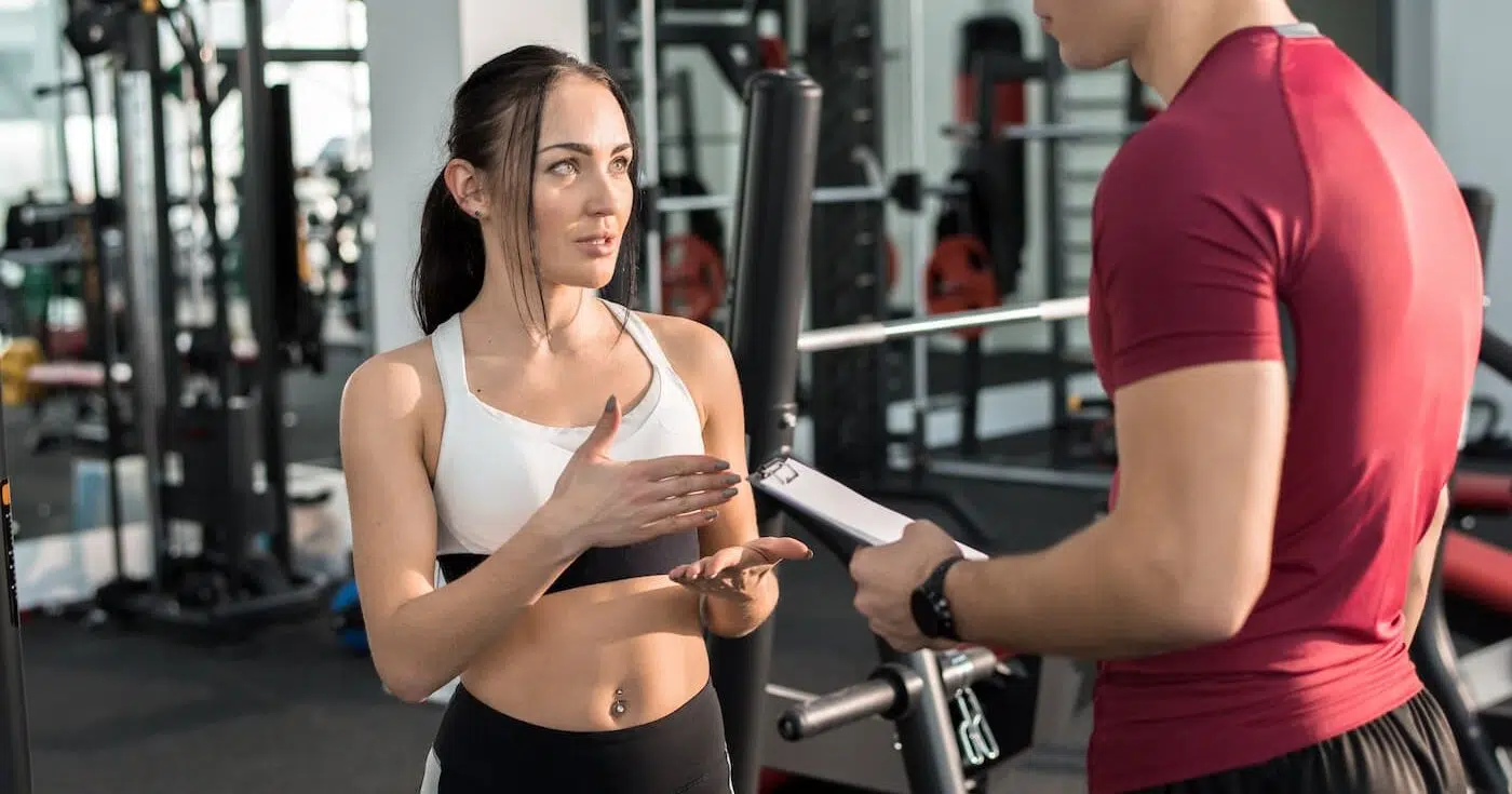 Young woman talking to personal fitness coach in modern gym e1645028424321