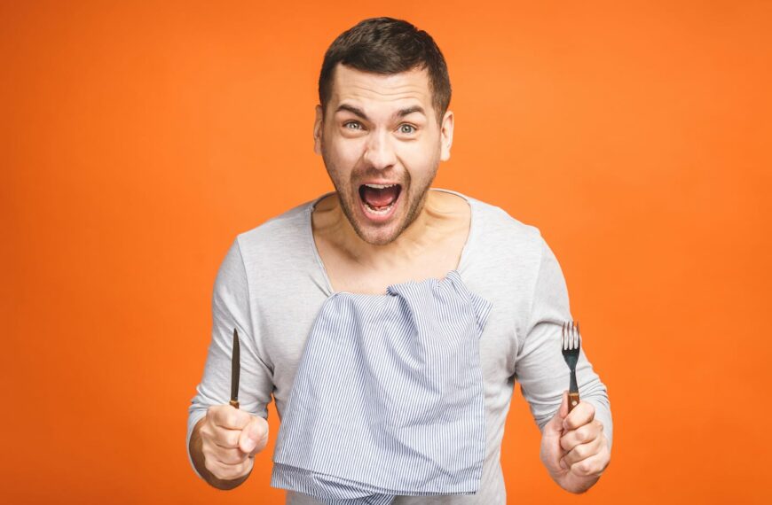 Young hungry crazy man holding a fork and a knife. Isolated on orange background