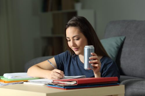 young person sits writing while holding a can of drink scaled