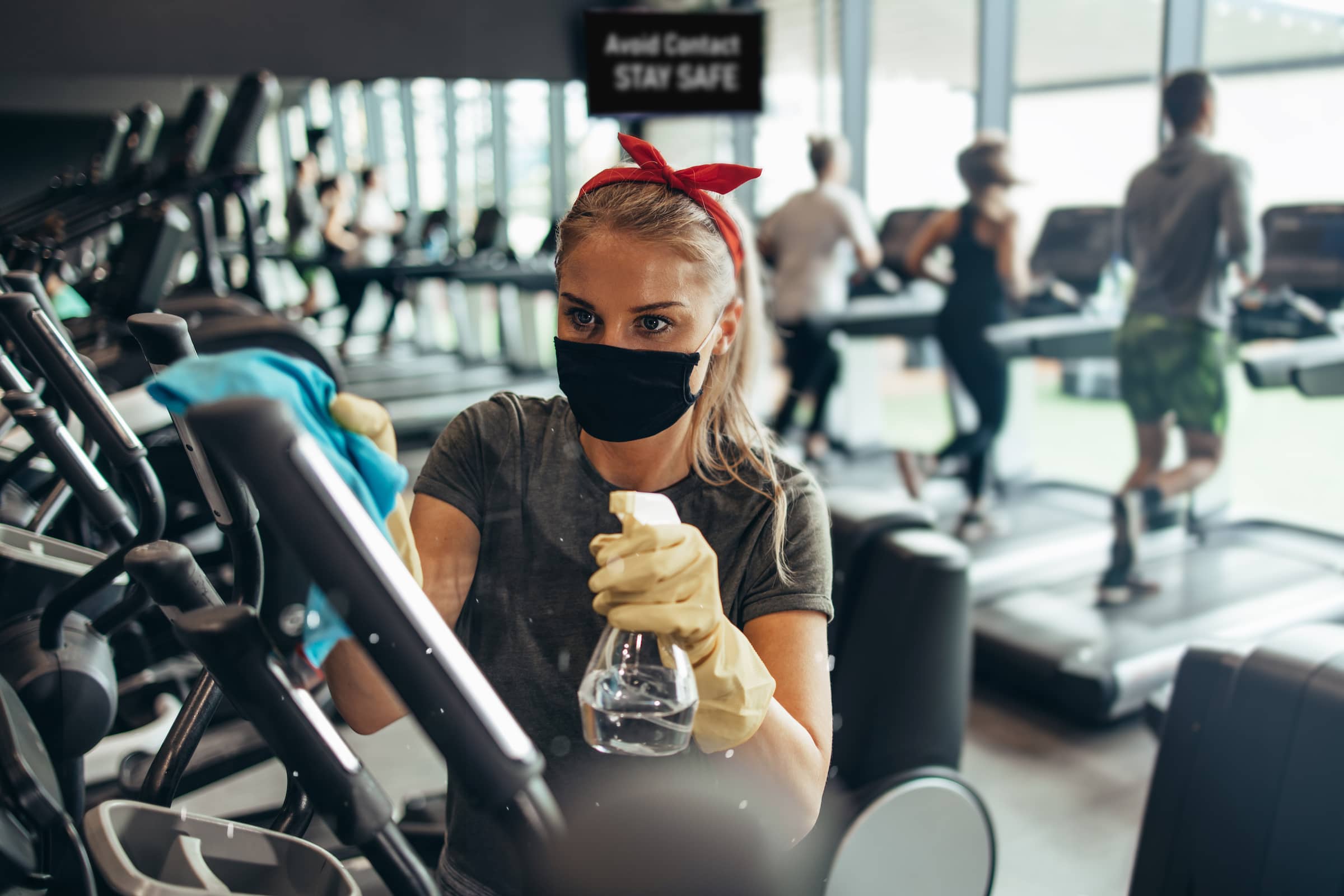 Young female worker disinfecting cleaning and weeping expensive fitness gym equipment with alcohol sprayer and cloth