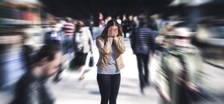 woman holds hands to head with blurred crowd around them e1648994268525