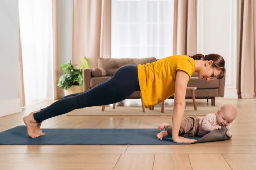 yogi mom smiling at her baby while doing push up on exercising mat at home