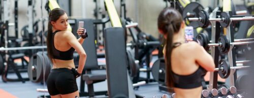 woman with smartphone taking mirror selfie in gym