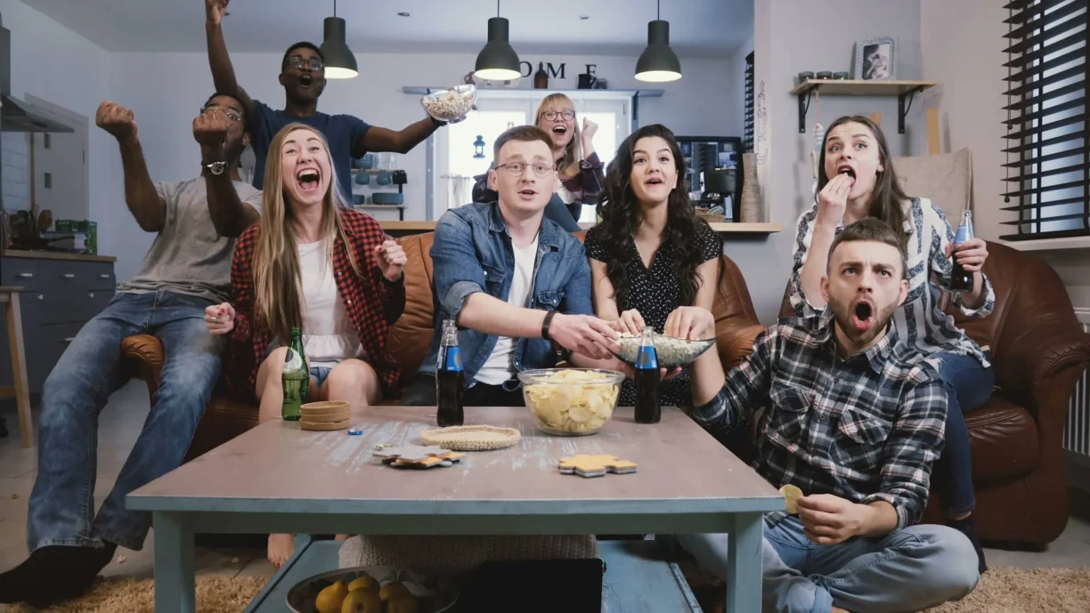 Friends watch sports on TV, cheer and celebrate. Happy diverse supporters fans sit on couch with popcorn and drinks.
