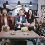 Friends watch sports on TV, cheer and celebrate. Happy diverse supporters fans sit on couch with popcorn and drinks.