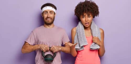 Photo of puzzled man holds weight, dressed in t shirt and white headband, his ethnic curly woman stands near, has towel around neck for wiping sweat, have workout together in fitness centre