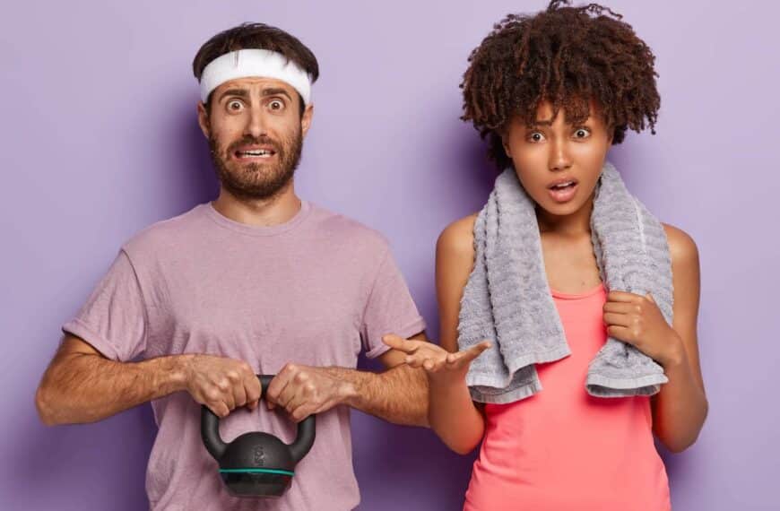 Photo of puzzled man holds weight, dressed in t shirt and white headband, his ethnic curly woman stands near, has towel around neck for wiping sweat, have workout together in fitness centre
