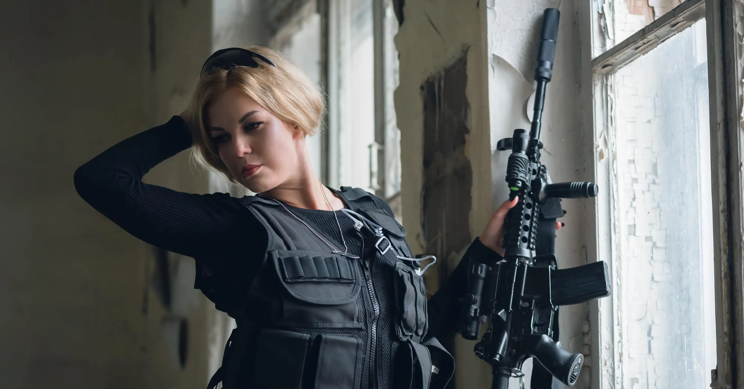 Young woman with airsoft rifle is posing near broken window.