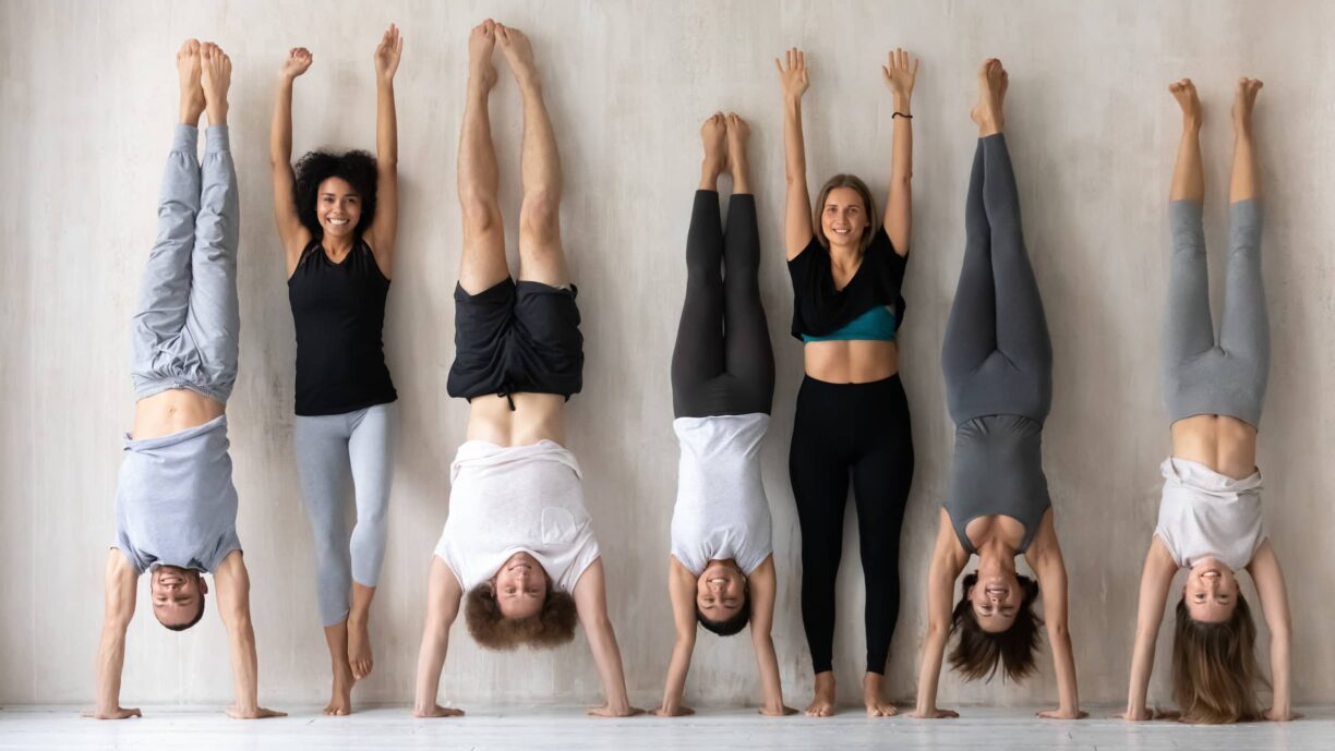 Happy young women in activewear standing near wall with raised arms while sportive mixed race people performing handstands