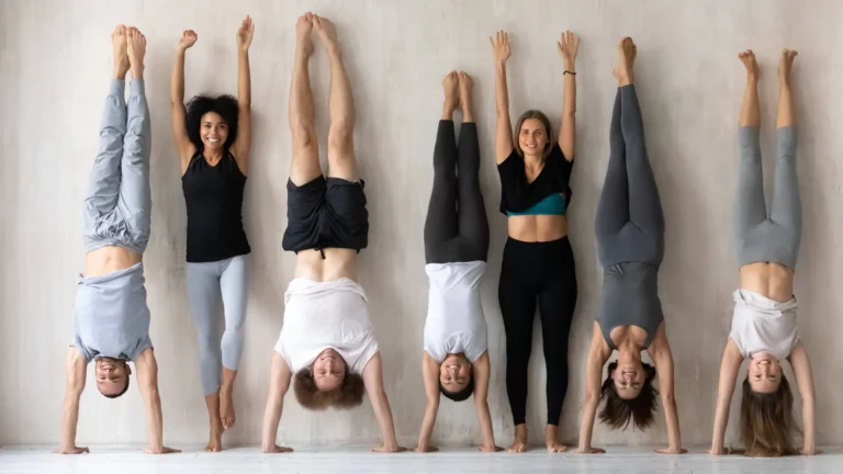Happy-young-women-in-activewear-standing-near-wall-with-raised-arms-while-sportive-mixed-race-people-performing-handstands