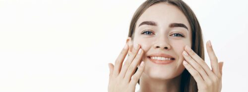 charming-relaxed-gentle-young-woman-making-cosmetological-procedure-applying-facial-cream-face-with-fingers-smiling-broadly-feeling-perfect-taking-care-skin