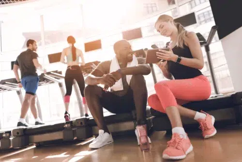 Athletes engage in modern treadmills in the bright gym. A man and a woman are sitting and talking next to the treadmills.
