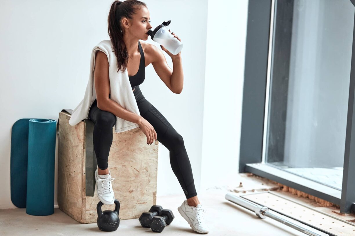 Young woman in leggings with towel on shoulders drinking water after fitness training