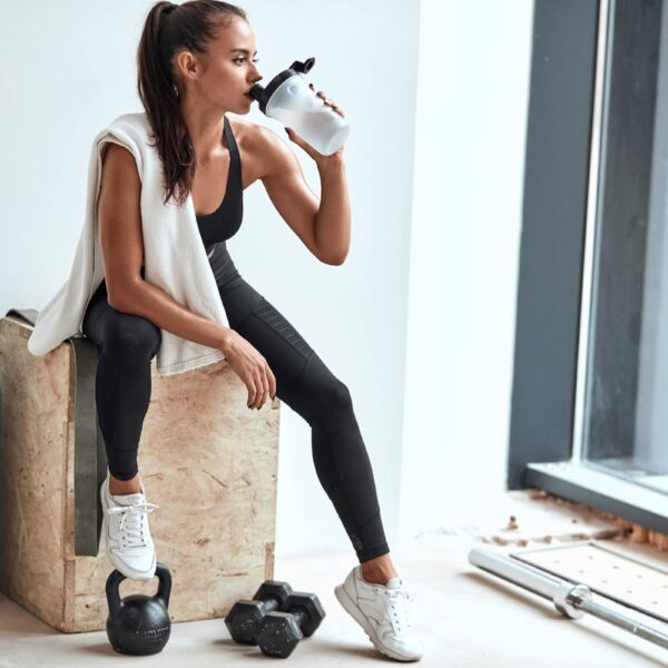 Young woman in leggins with towel on shoulders drinking water after fitness training