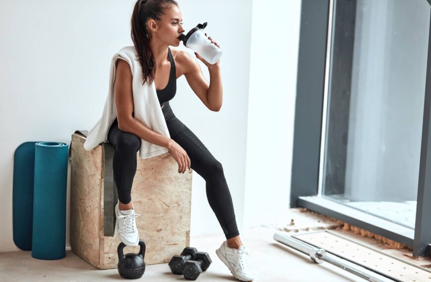 Young woman in leggings with towel on shoulders drinking water after fitness training