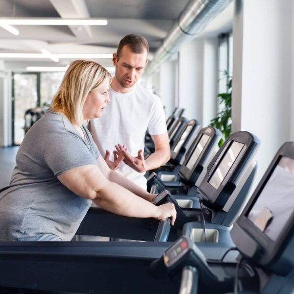 Overweight woman exercising on treadmill in gym. Personal trainer helping her.