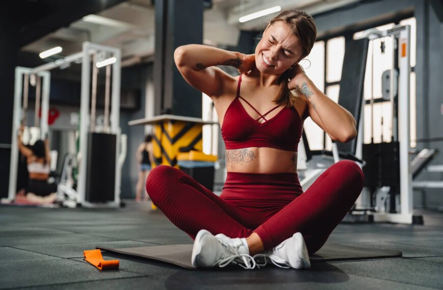 Unhappy athletic sportswoman with neck pain sitting on mat in gym