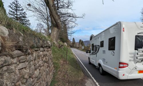 A road with a caravan driving down it and little space for anyone to walk safely