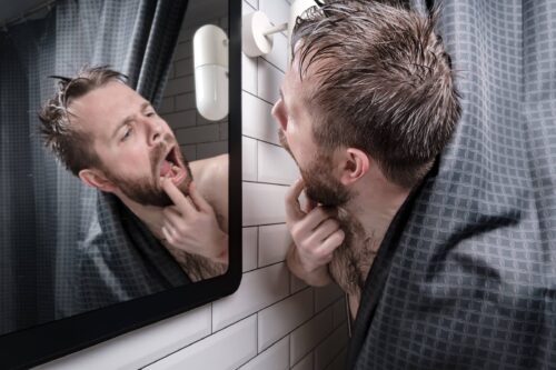 Man carefully examines his teeth in the mirror, peering out from behind the shower curtain.