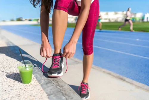 Woman runner tying running shoes drinking green smoothie cup juice drink before race