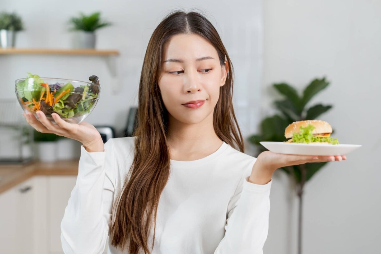 Young woman, girl confused choose, choice eating food, hand holding bowl of vegetable salad and hamburger