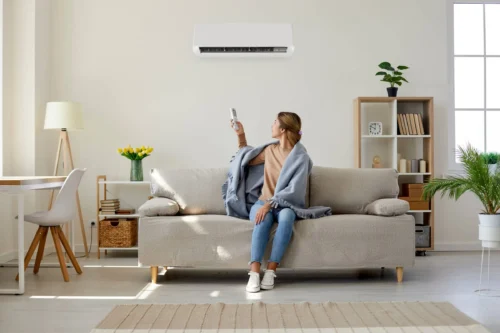 Woman who's sitting on sofa under warm plaid in living room switches off her air conditioner on wall. Young girl adjusting modern AC system