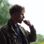 Casual thoughtful male smoking electronic cigarette while standing outdoors in park looking away