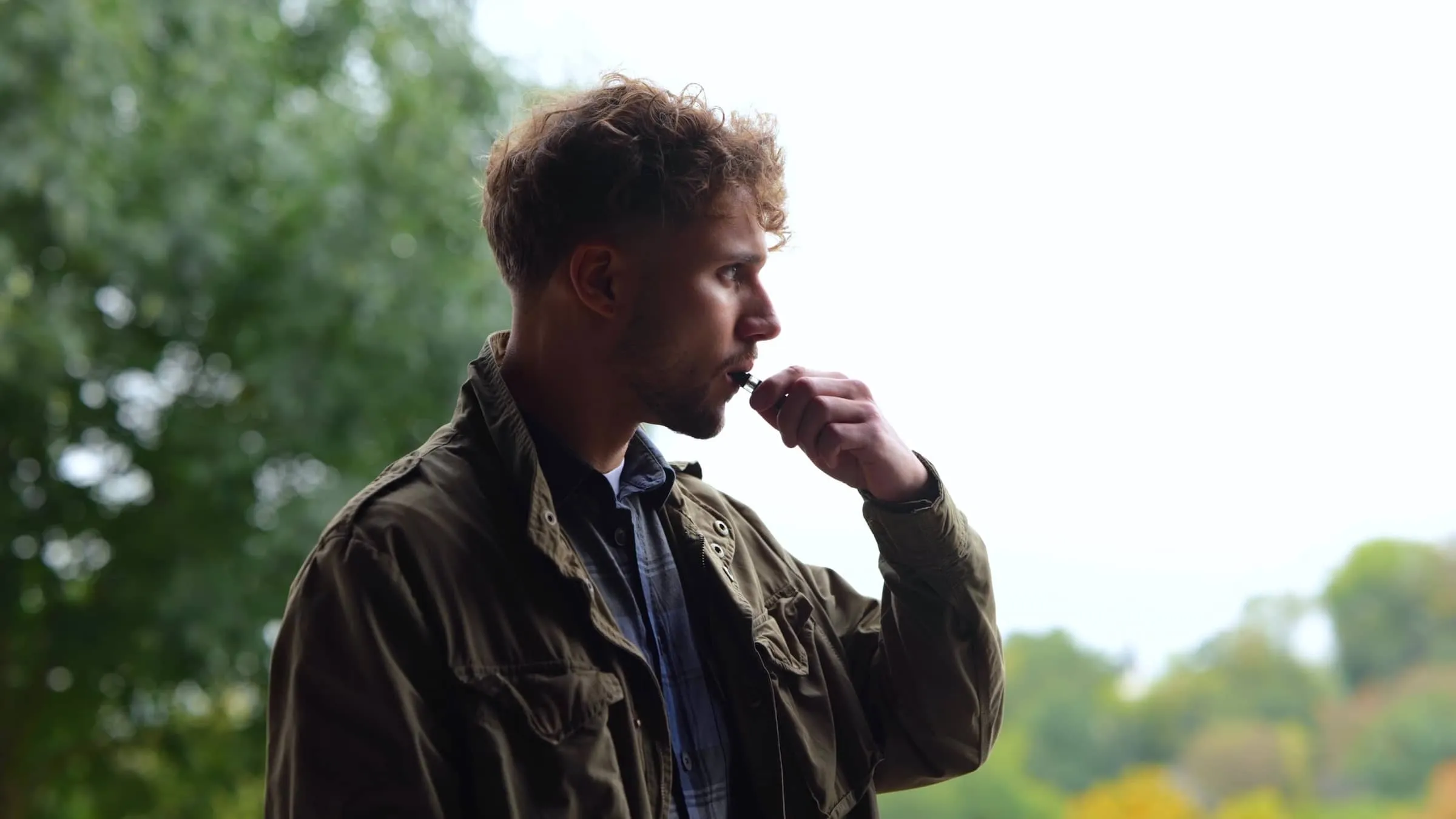 Casual thoughtful male smoking electronic cigarette while standing outdoors in park looking away
