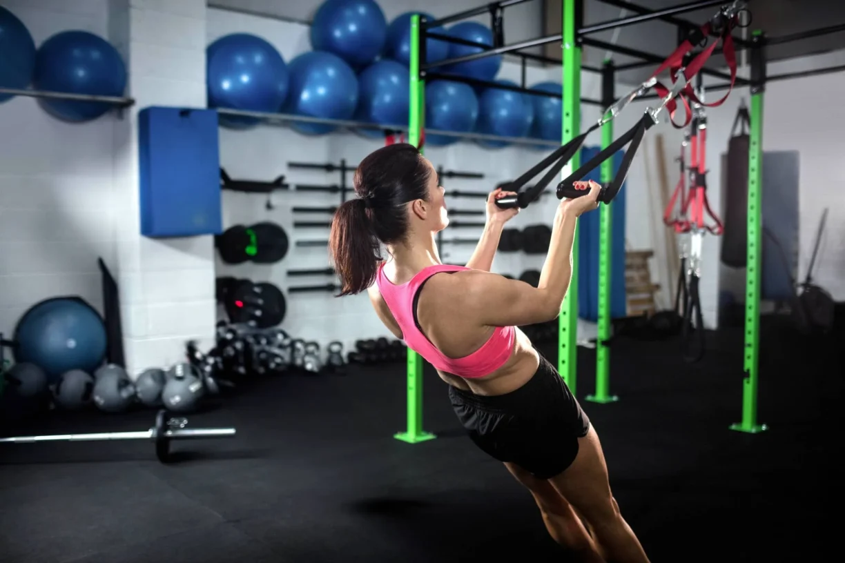 Female fitness trainee gripping black suspension straps and pulling body at gym functional rig area