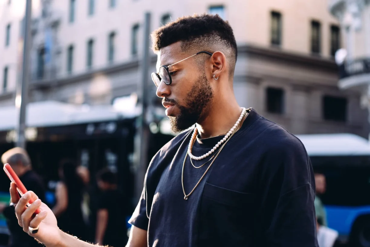 Stylish man in his 20s with beard and glasses using smartphone in urban setting