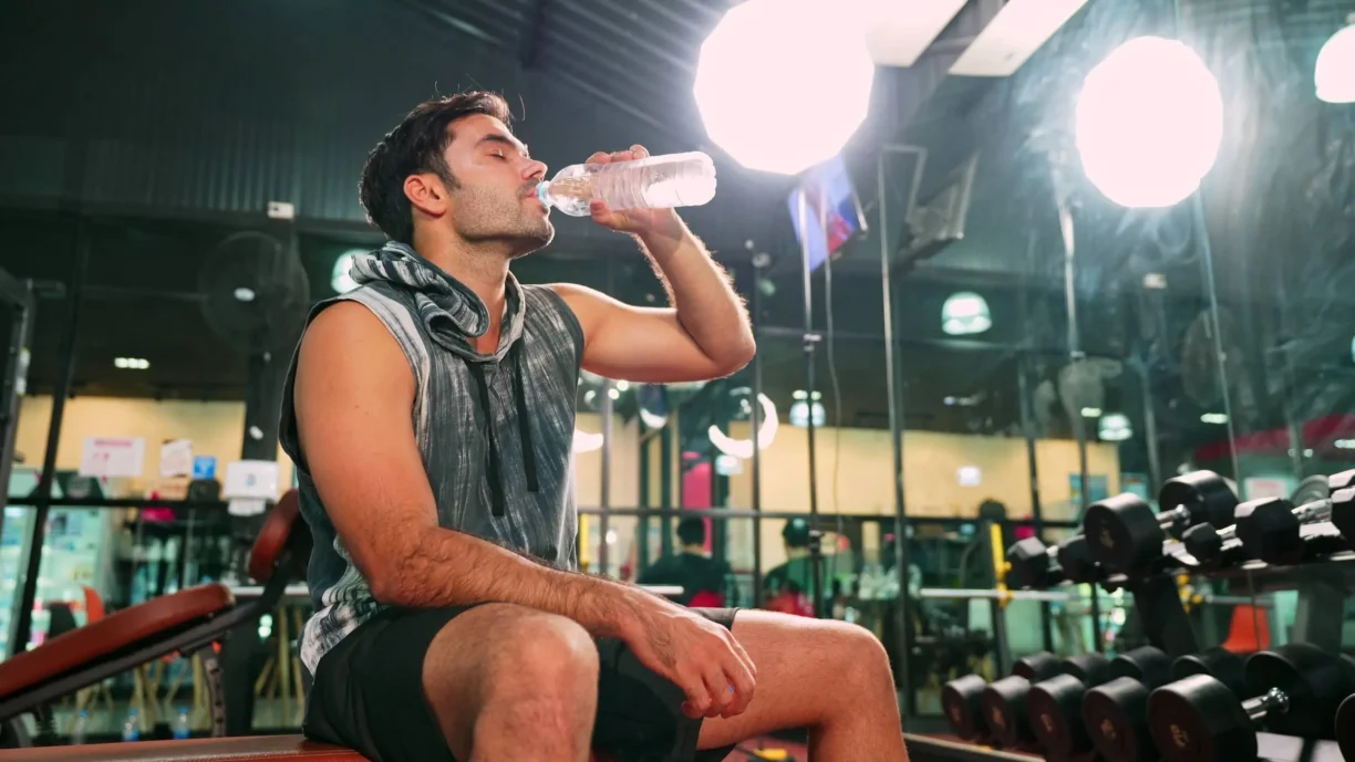 male bodybuilder is sitting on fitness bench and drinking water to cool down his body after workout in gym