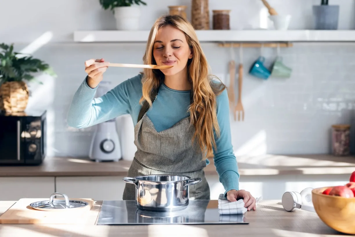 Woman cooking healthy food in casserole while blowing the spoon to taste the food