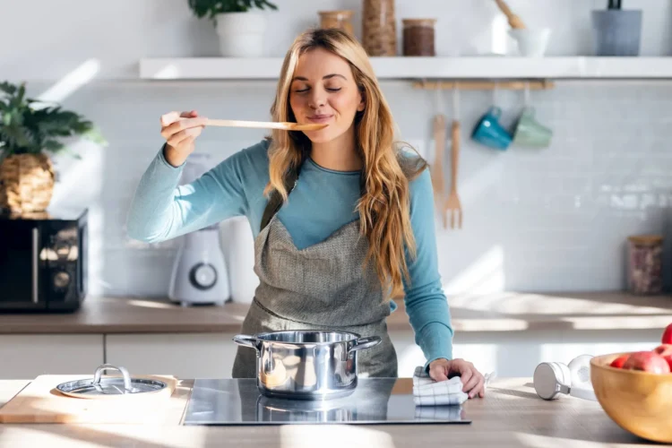 Woman cooking healthy food in casserole while blowing the spoon to taste the food