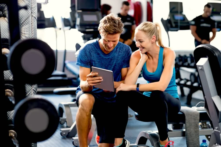 Smiling personal trainer using digital tablet while talking to blonde women at gym