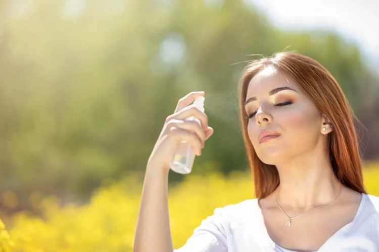 Young woman spraying mist on face with eyes closed in that moment she is on blooming flowers field