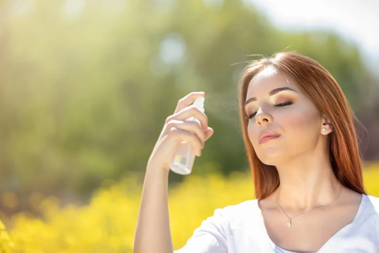 Young woman spraying mist on face with eyes closed in that moment she is on blooming flowers field